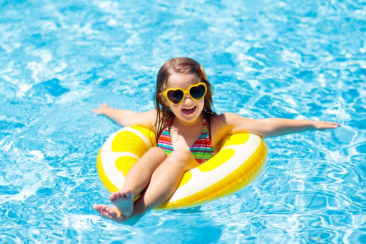 Smiling child in heart-shaped sunglasses, floating on a yellow inflatable ring in a bright blue swimming pool with arms outstretched in mesa az