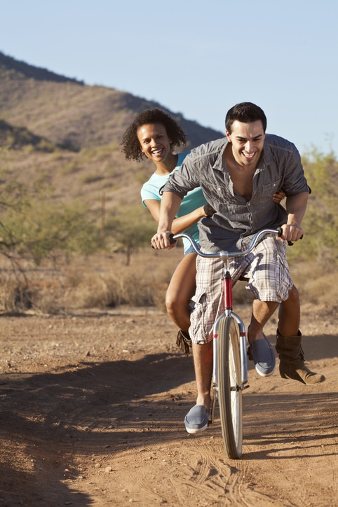 Smiling couple on a bicycle ride, with the man pedaling and the woman holding onto him, on a dirt path in a desert landscape in mesa az