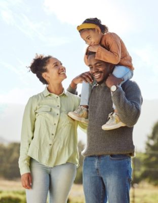 Smiling family outdoors with a man carrying a young child on his shoulders and a woman walking beside them in mesa az