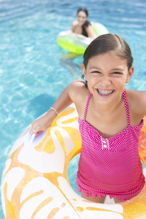 Smiling young girl with braces in a pink swimsuit, holding onto a yellow pool float in a swimming pool, with another child in the background in mesa az