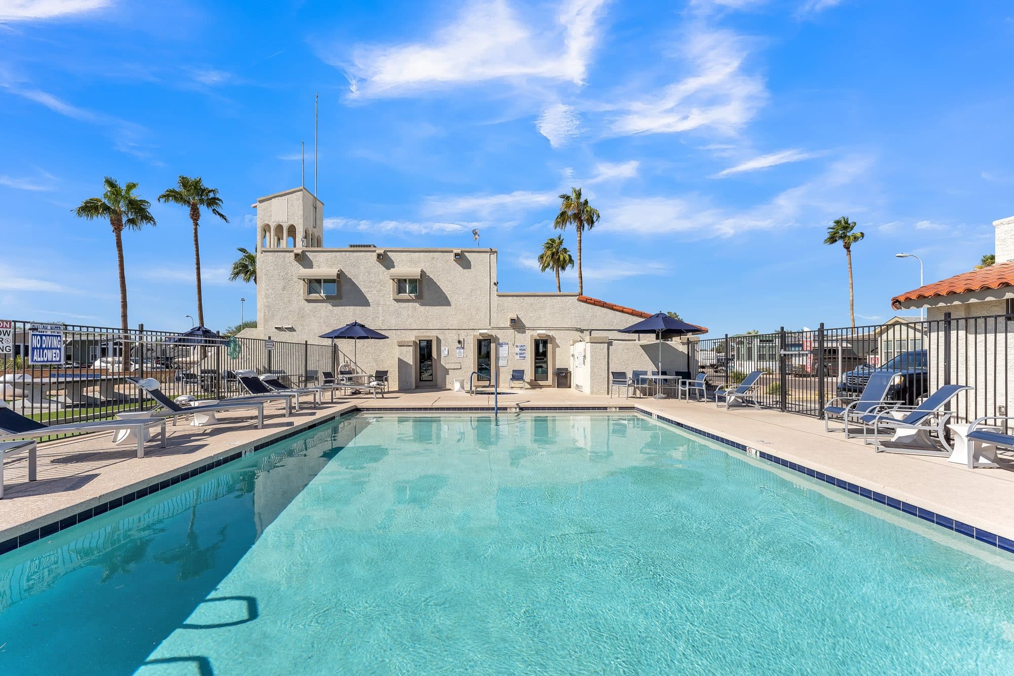 Outdoor swimming pool area surrounded by lounge chairs, a beige building with a tower, and palm trees under a bright blue sky in mesa az